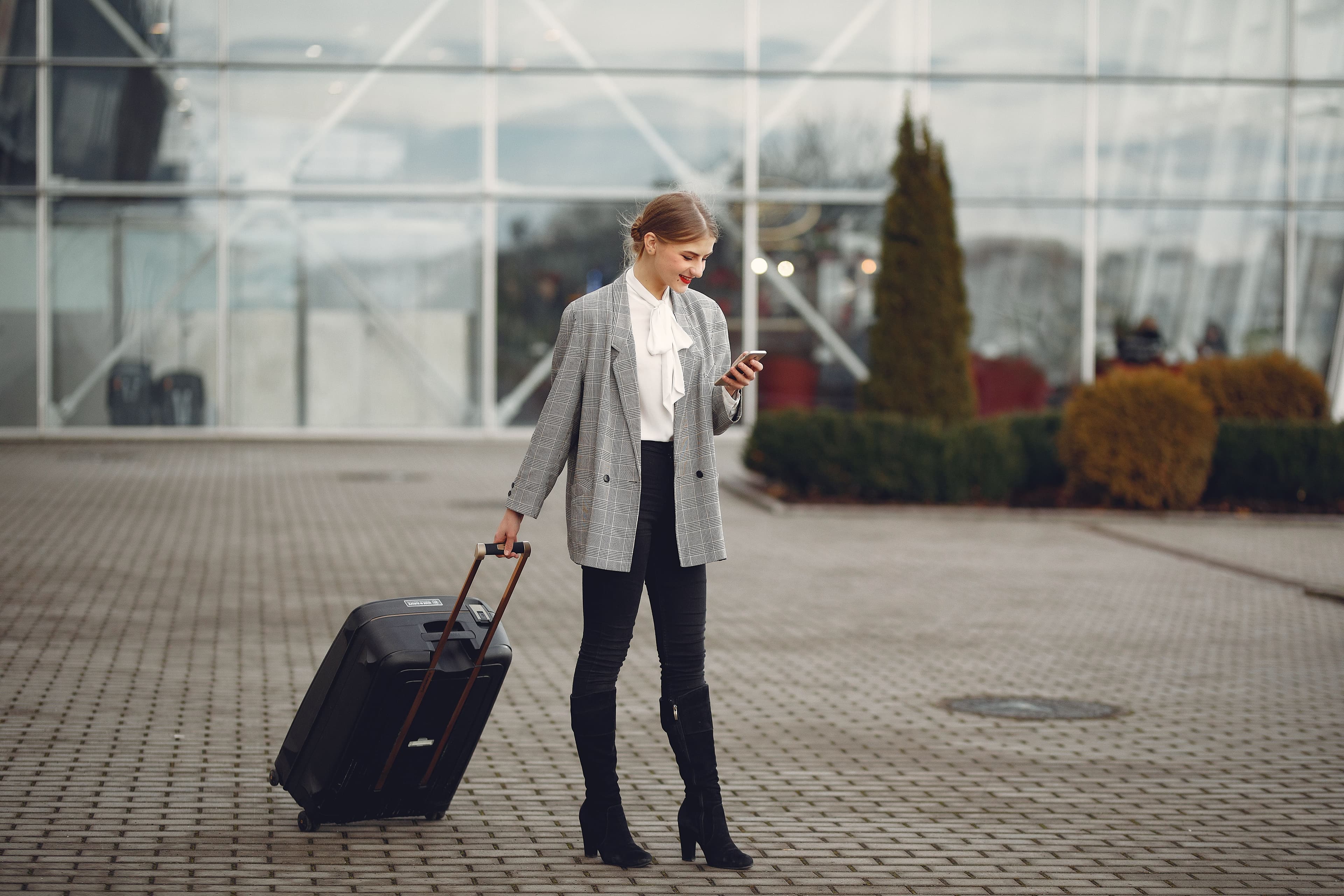 Business traveler with a suitcase checking her phone outside a modern airport